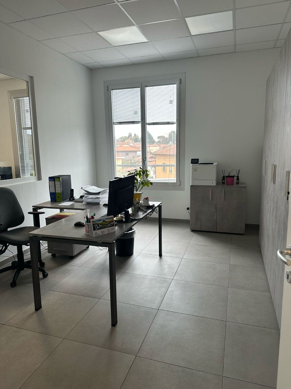 Piccolo ufficio con scrivania, monitor, sedia e parete finestrata in una stanza luminosa piastrellata.