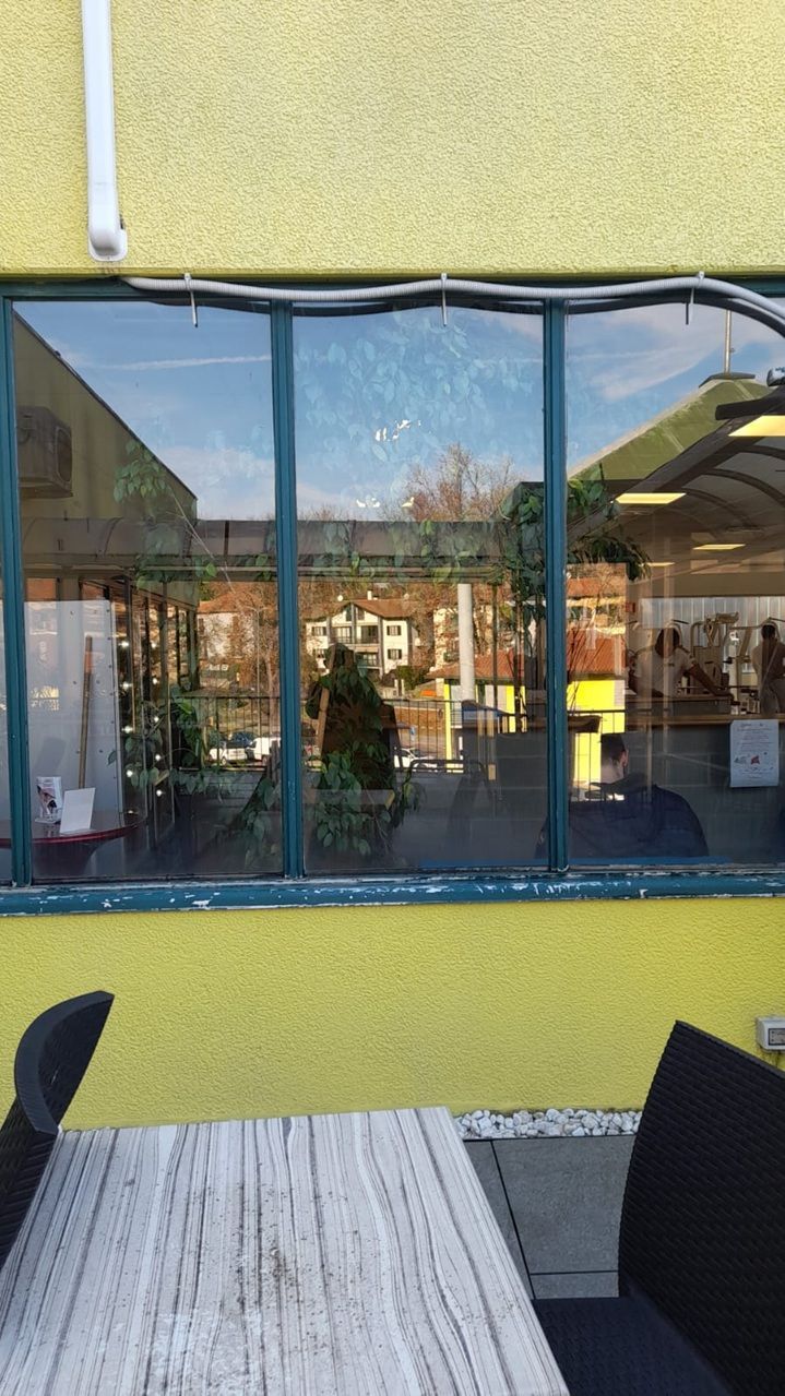 Tavolino da caffè all'aperto di fronte a una finestra con cornice blu su un edificio giallo, che riflette gli alberi e le case vicine.