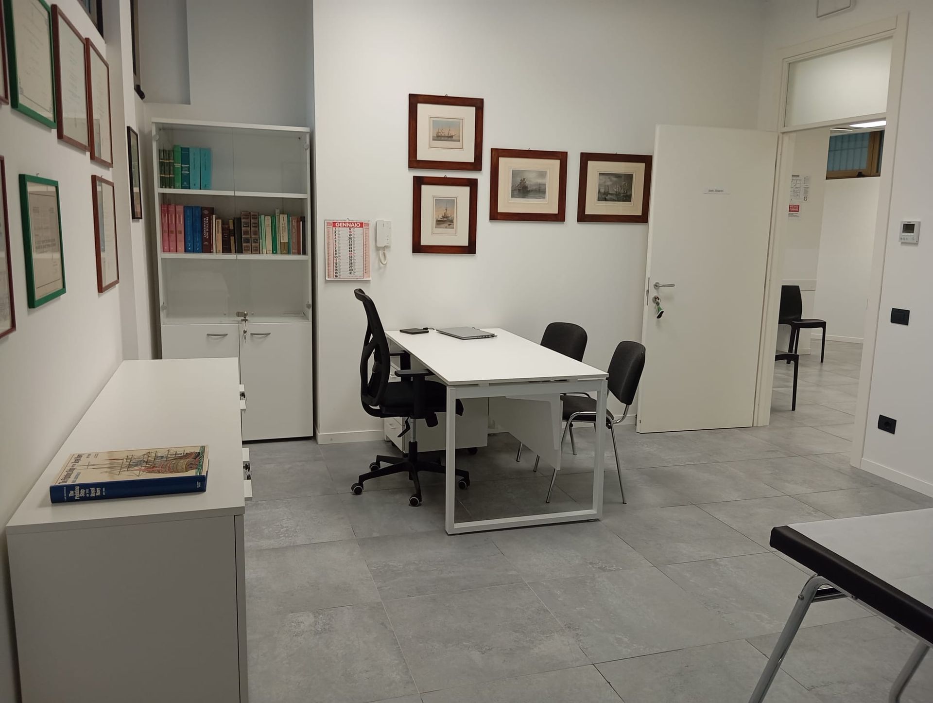 Sala riunioni in ufficio con scrivania bianca, sedia nera, quadri alle pareti e pavimento in piastrelle grigie.