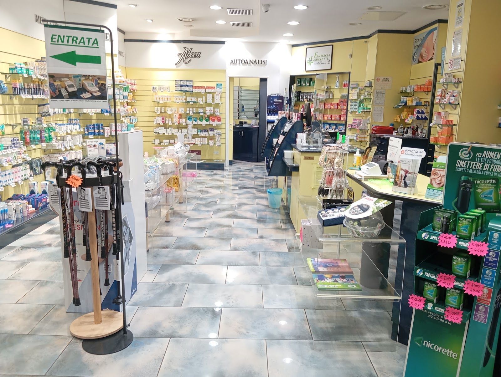 Corridoio della farmacia con scaffali pieni di medicinali, espositori e una persona vicino al bancone sul retro.