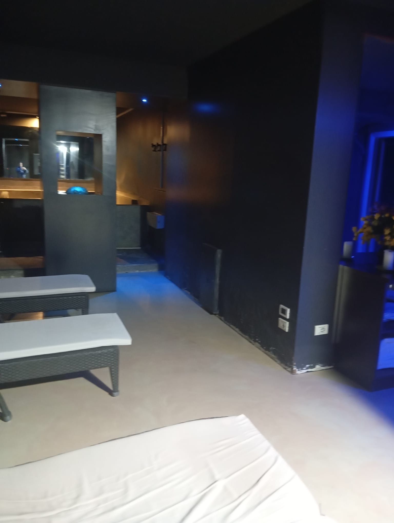Camera d'albergo poco illuminata con due letti, pareti scure, una panca e luci blu soffuse vicino a una finestra.