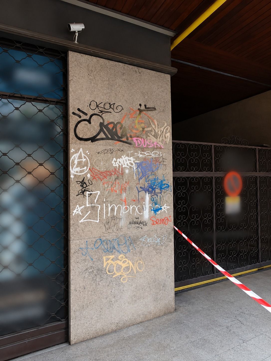 Un pilastro di cemento imbrattato di graffiti accanto a un cancello di metallo, con nastro di segnalazione rosso e bianco e un cartello di divieto di sosta sotto un passaggio coperto.