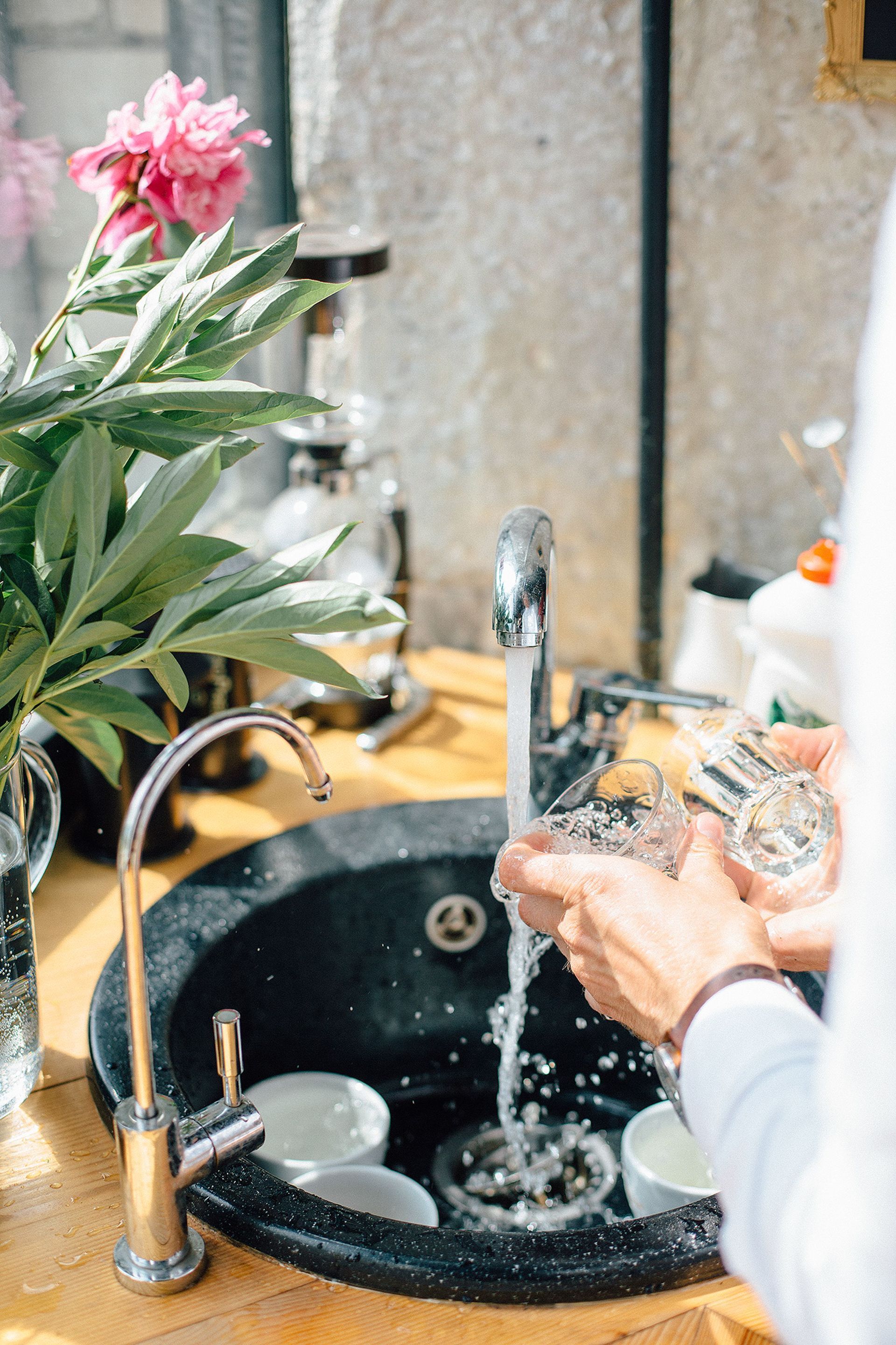 Hands washing dishes in a black sink with running water beside a vase of pink flowers