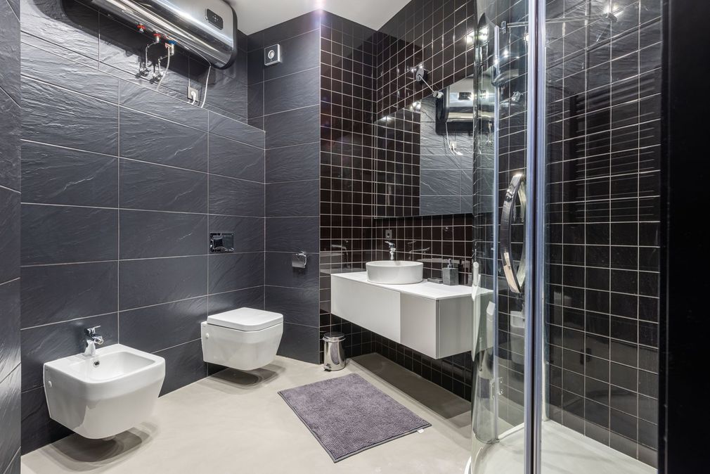 Modern black-tiled bathroom with sink, toilet, bidet, shower, and purple bath mat