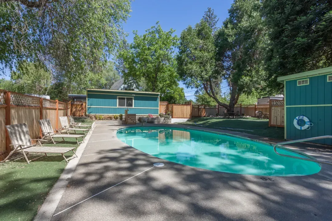 Backyard with a turquoise swimming pool, patio, wooden fence, and trees on a sunny day