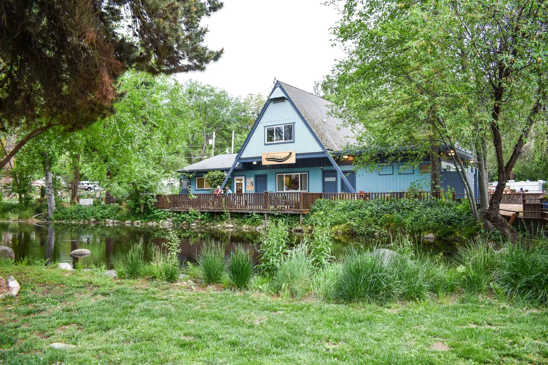 Blue house with a steep roof and deck, surrounded by lush green trees and grass.