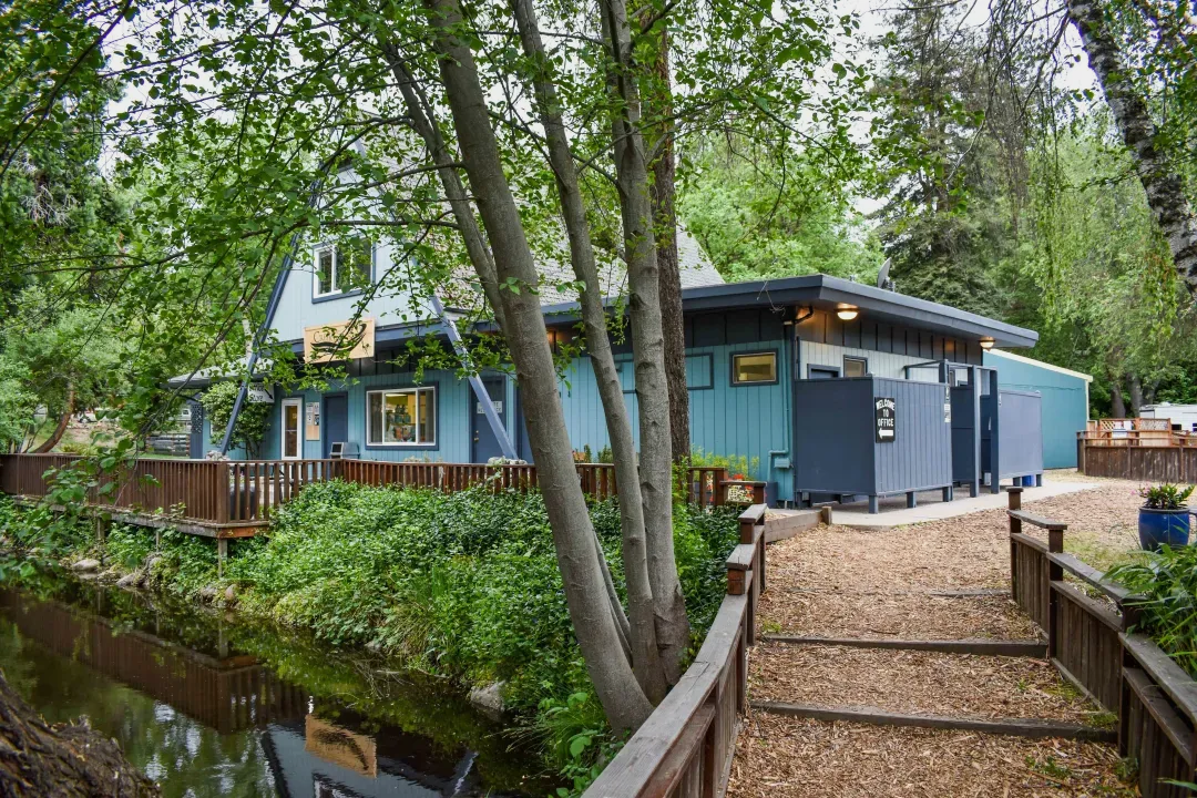 Blue building beside a wooded stream with a gravel path and wooden bridge railing