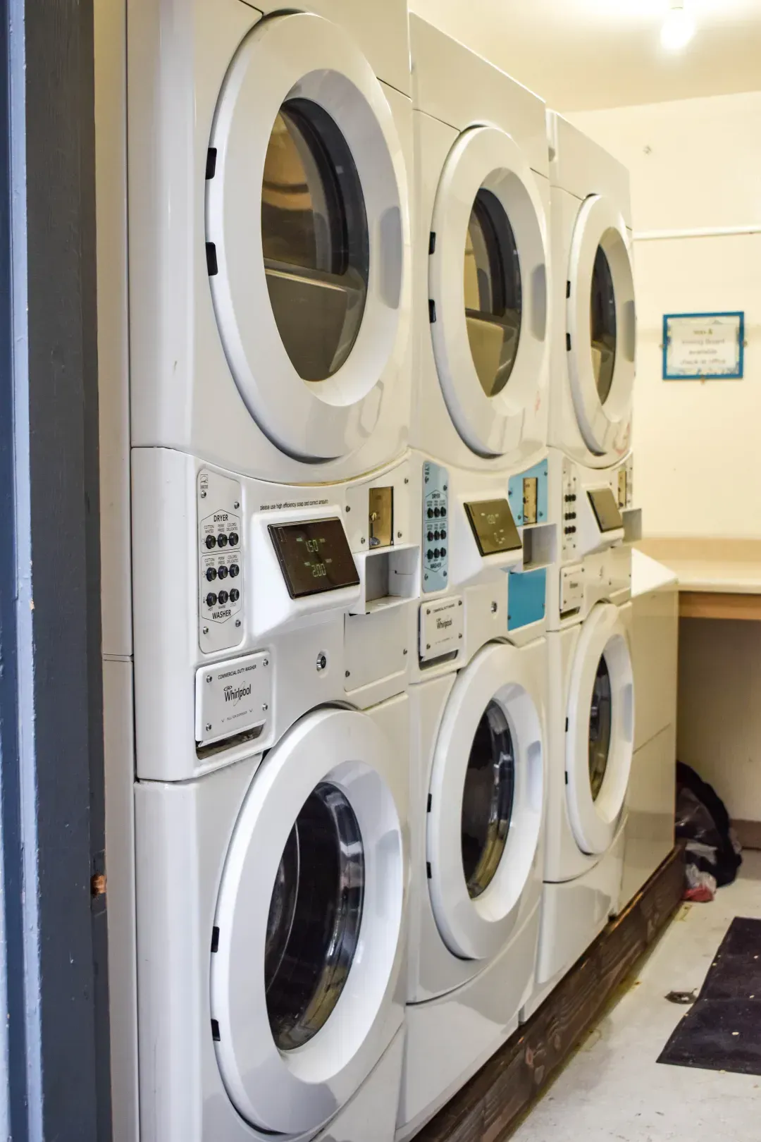 Stacked white washing machines and dryers in a laundromat, with a counter and dark mats along the wall.