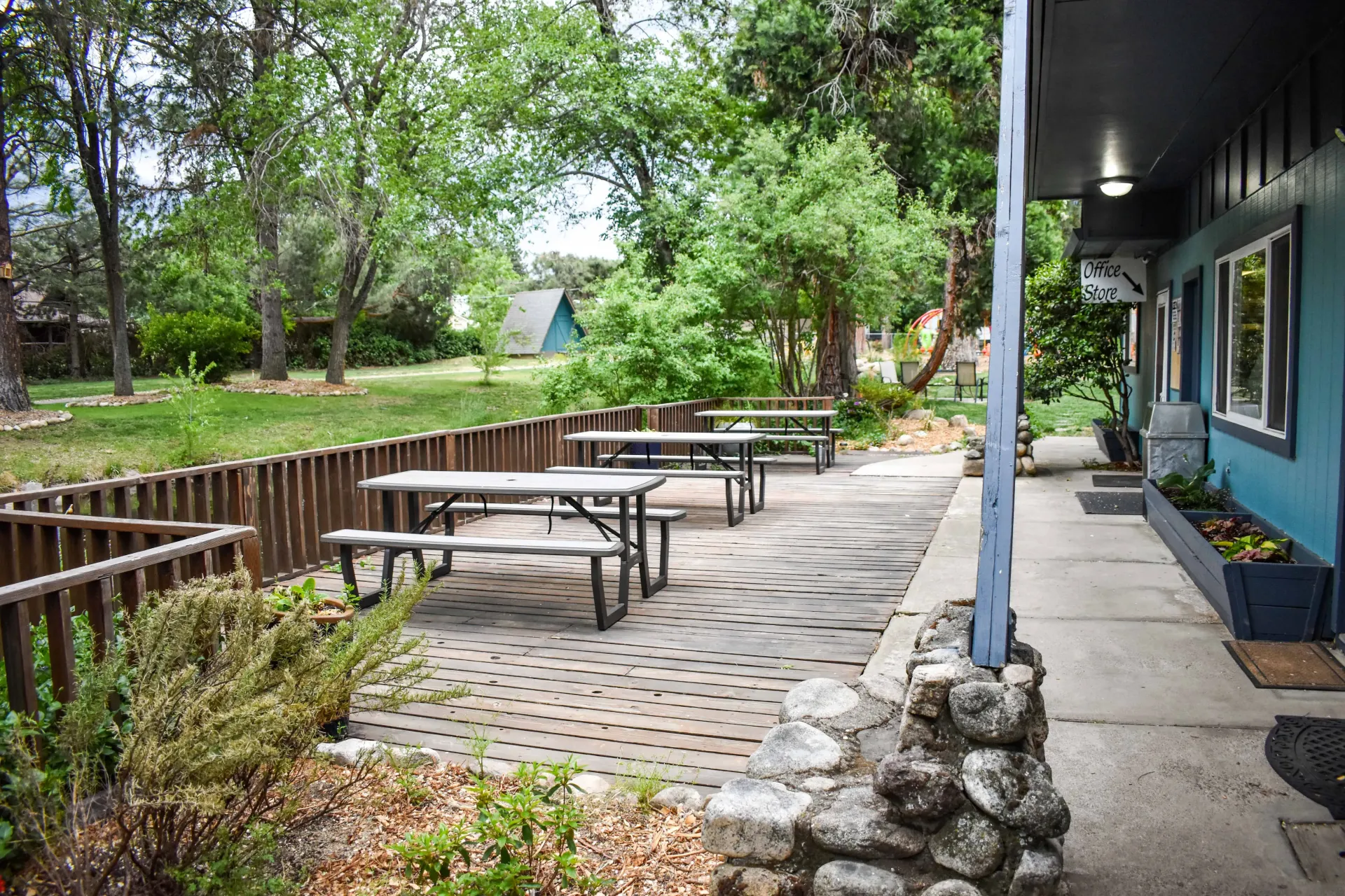 Outdoor patio with picnic tables beside a blue building, surrounded by trees and greenery
