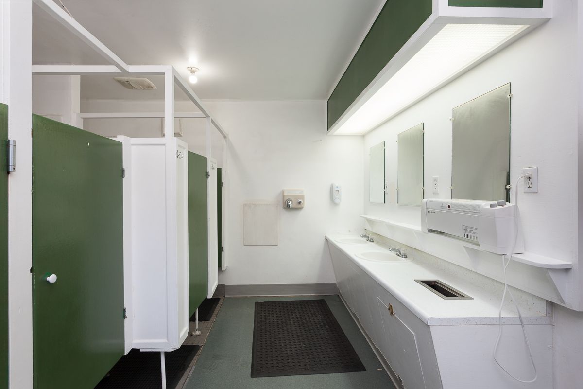 Bright clean restroom with green stall doors, white sinks, mirrors, and a black floor mat.