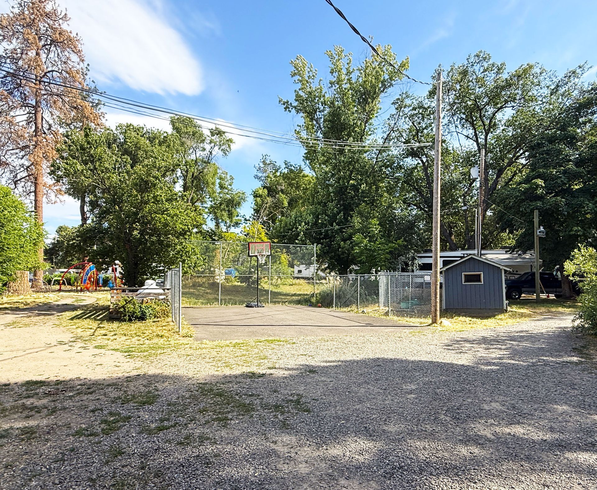 Gravel driveway leading to a fenced yard with trees and a small gray shed under a blue sky