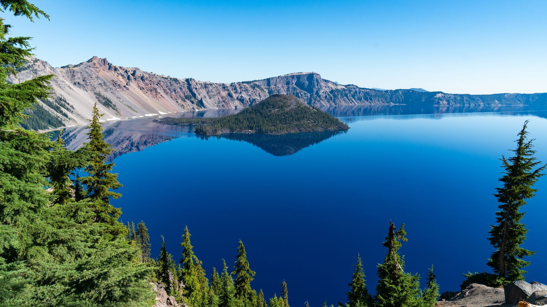 Blue crater lake surrounded by rocky cliffs and evergreen trees under a clear sky