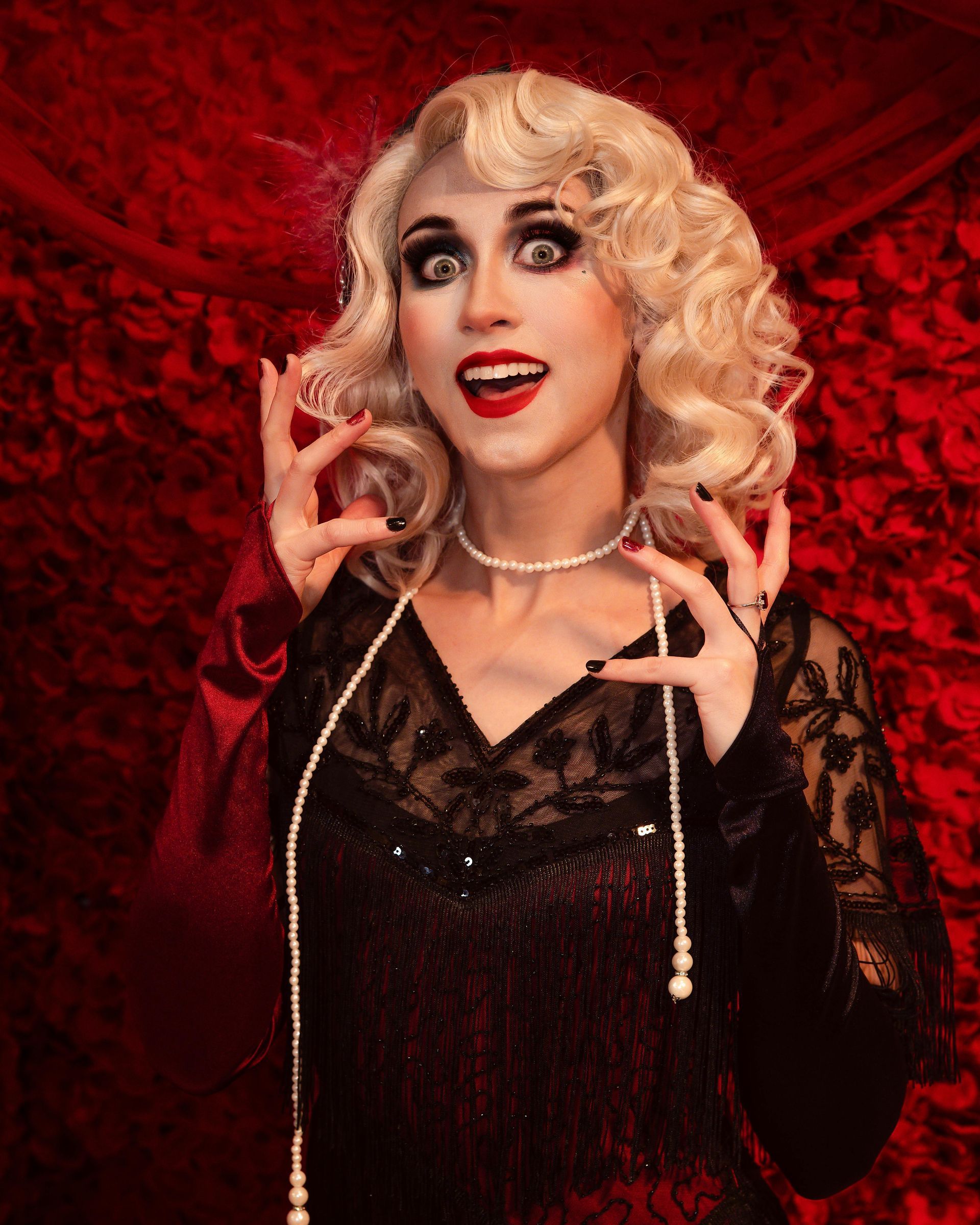 Woman with blonde curls and red lipstick posing against a red textured backdrop, wearing a black lace dress.