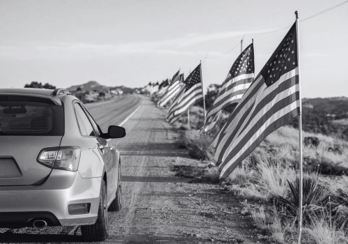 A car is parked beside a rural road lined with American flags stretching into the distance.