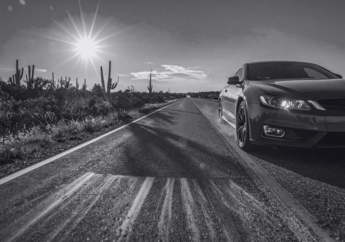 Black sedan driving on a desert highway under bright sunlight with cacti and open landscape.