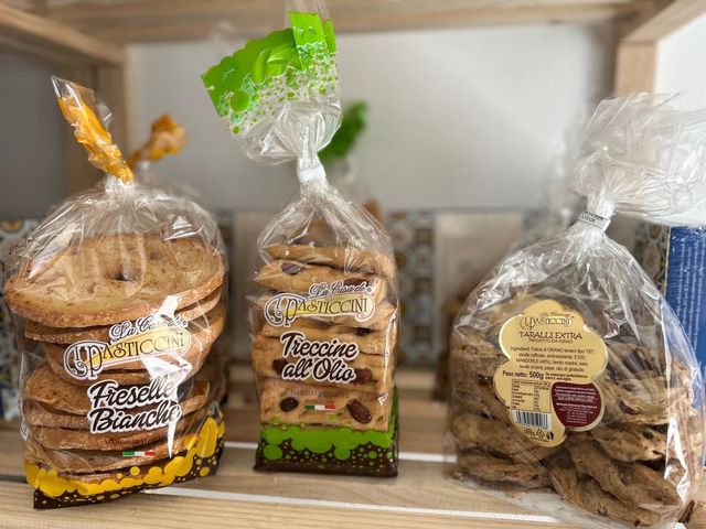 Biscotti assortiti confezionati in sacchetti trasparenti su uno scaffale, con etichette beige, verdi e marroni.