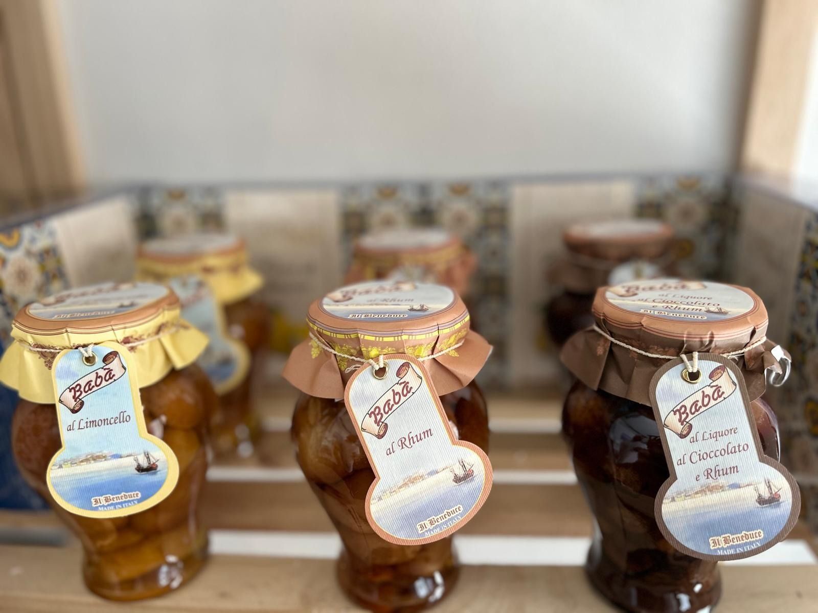Tre barattoli di conserve con coperchi dorati ed etichette decorative su uno scaffale di legno.