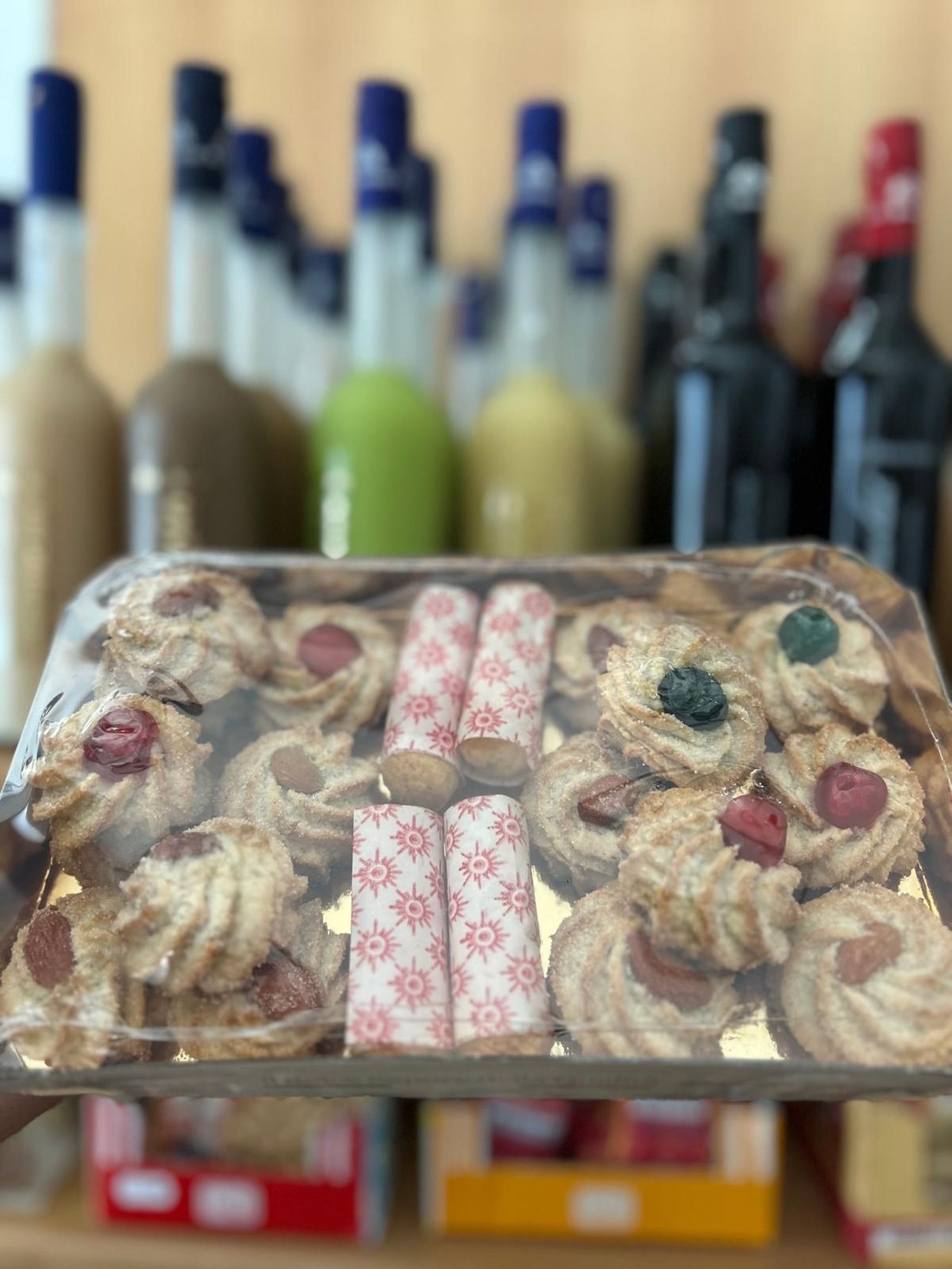 Biscotti assortiti in una scatola di plastica trasparente, con bottiglie di vino sfocate sullo sfondo.