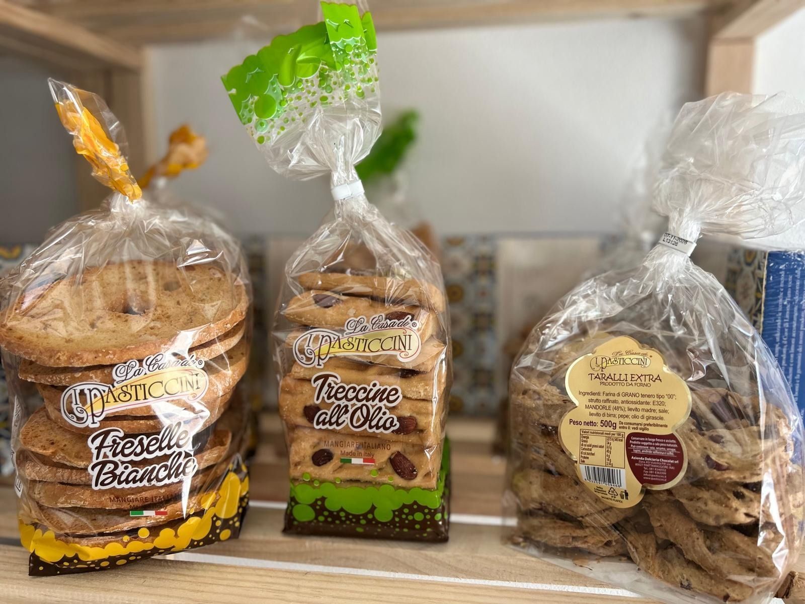 Su uno scaffale, tre sacchetti di biscotti assortiti, avvolti in plastica trasparente con etichette colorate.