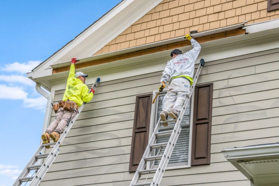 Two workers on ladders painting a house exterior with beige siding and brown shutters