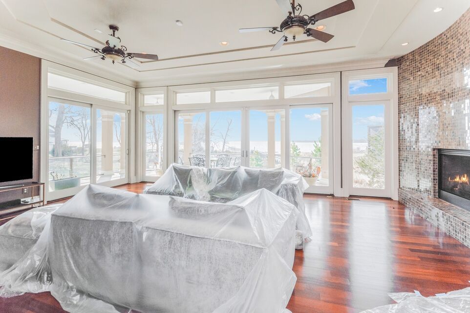 Bright living room with large windows, hardwood floors, and furniture wrapped in plastic during renovation