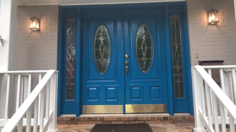 Blue double front doors with oval glass panels, flanked by wall lanterns and white porch railings.