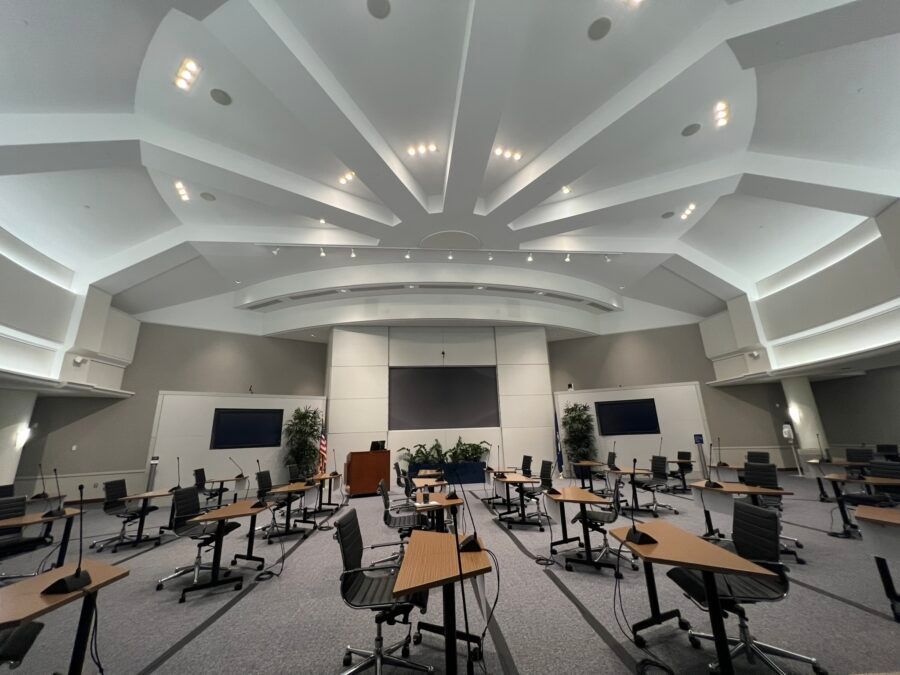Empty music rehearsal room with scattered chairs, music stands, and a stage under a bright vaulted ceiling