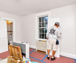 Worker painting a white wall in a small room with a radiator and doorway visible.
