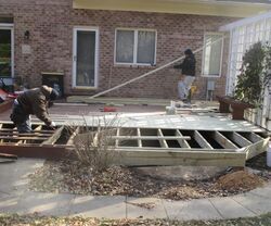 Two workers assembling a wooden deck frame in a backyard beside a brick house.