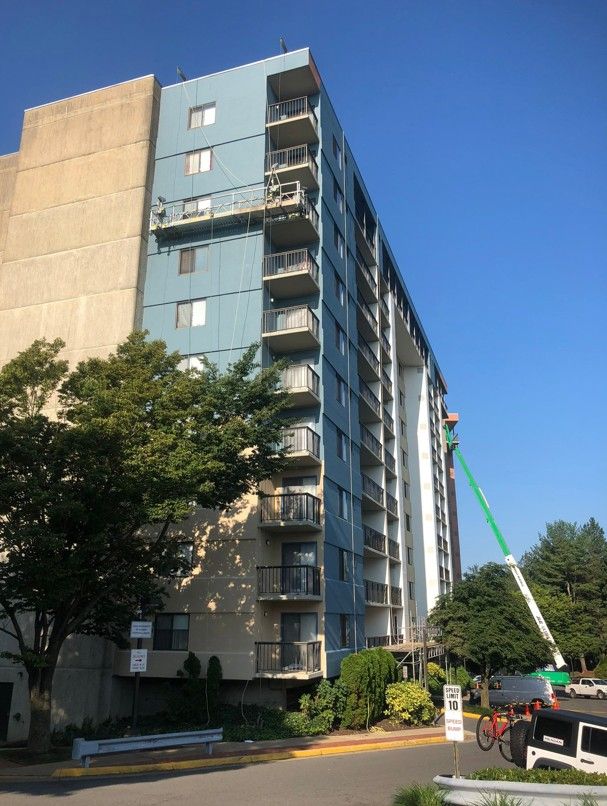 Tall blue apartment building with a crane beside it on a sunny street, partly shaded by trees