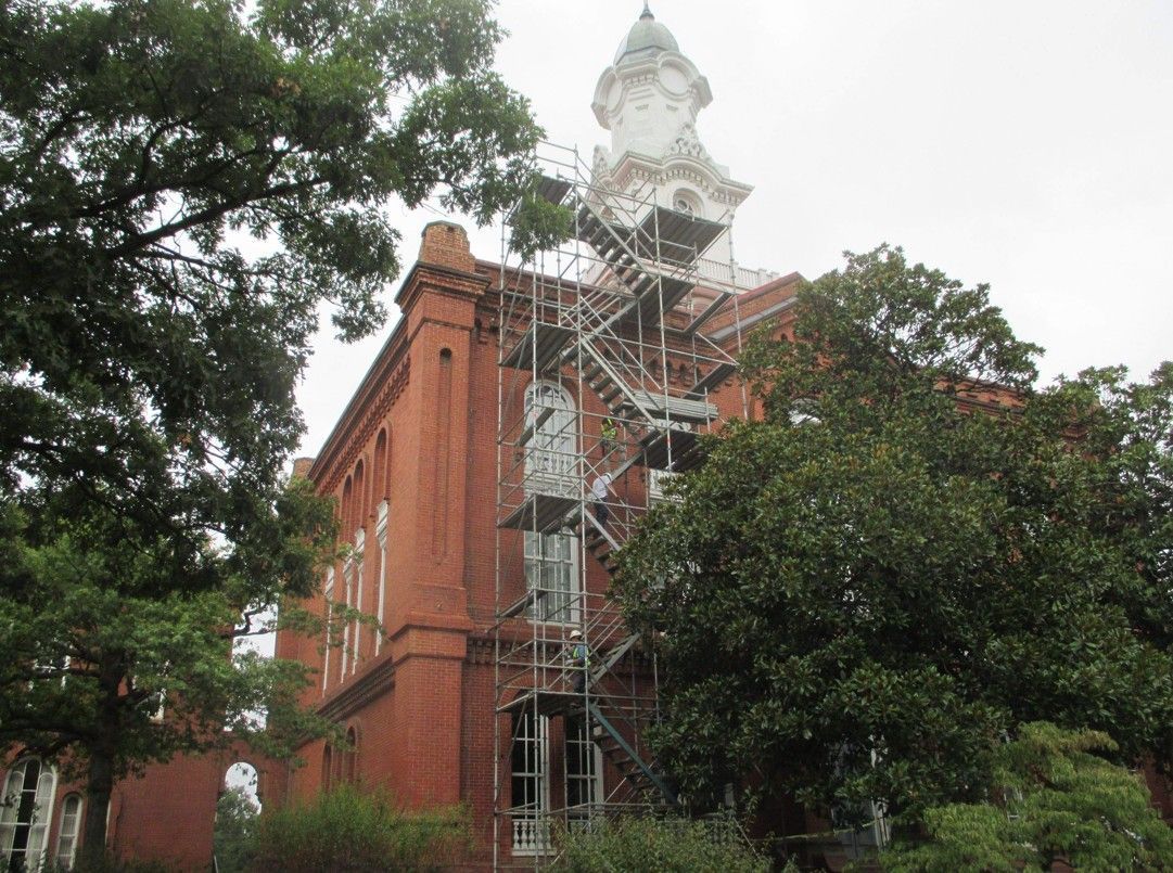 Red brick building with white clock tower under scaffolding, partly obscured by trees