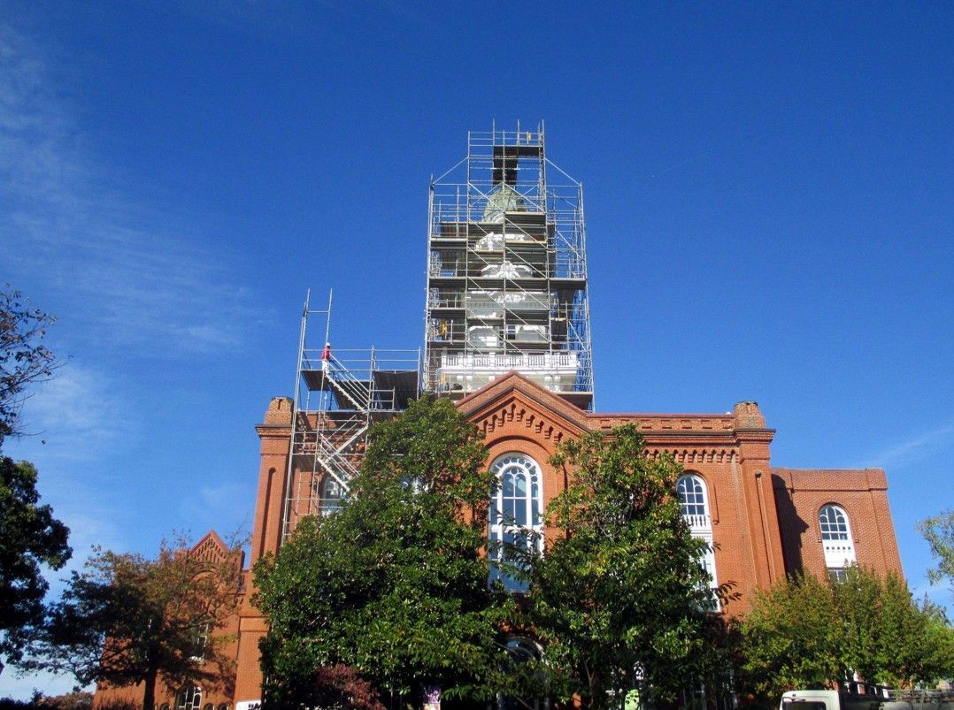 Red brick building with tower under scaffolding, framed by trees against a clear blue sky