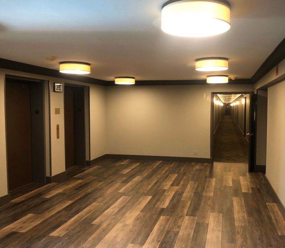 Empty modern lobby with wood-look flooring, beige walls, round ceiling lights, and a staircase in back.