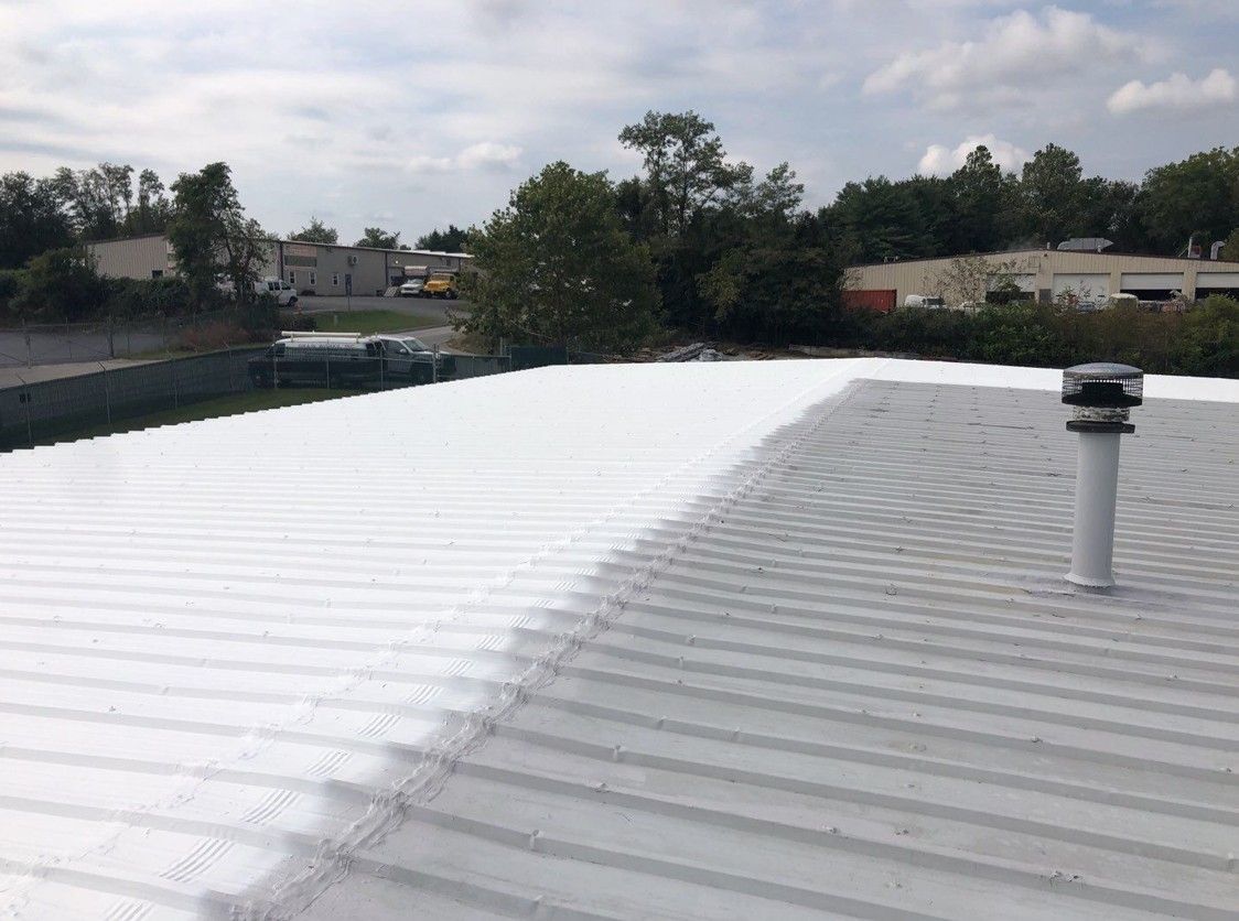 White metal rooftop under cloudy sky, with a vent pipe and trees in the distance