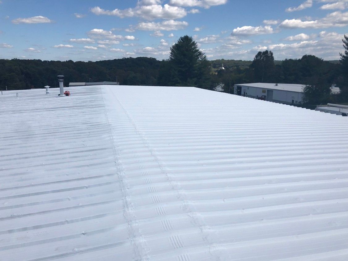 Wide white metal roof under a blue sky with clouds, trees and buildings in the distance