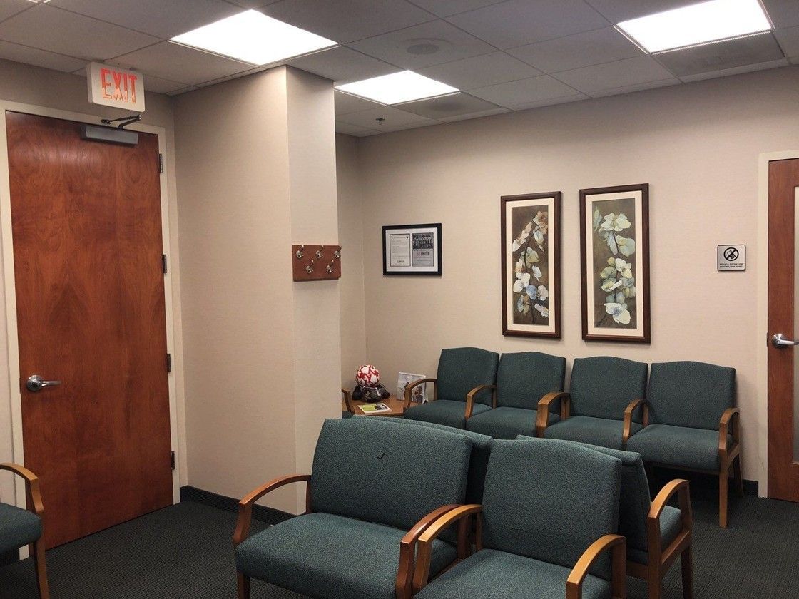 Empty waiting room with teal chairs, wooden door, framed wall art, and ceiling lights.