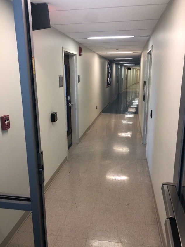 Long, empty office hallway with white walls, beige tile floor, and overhead fluorescent lights.