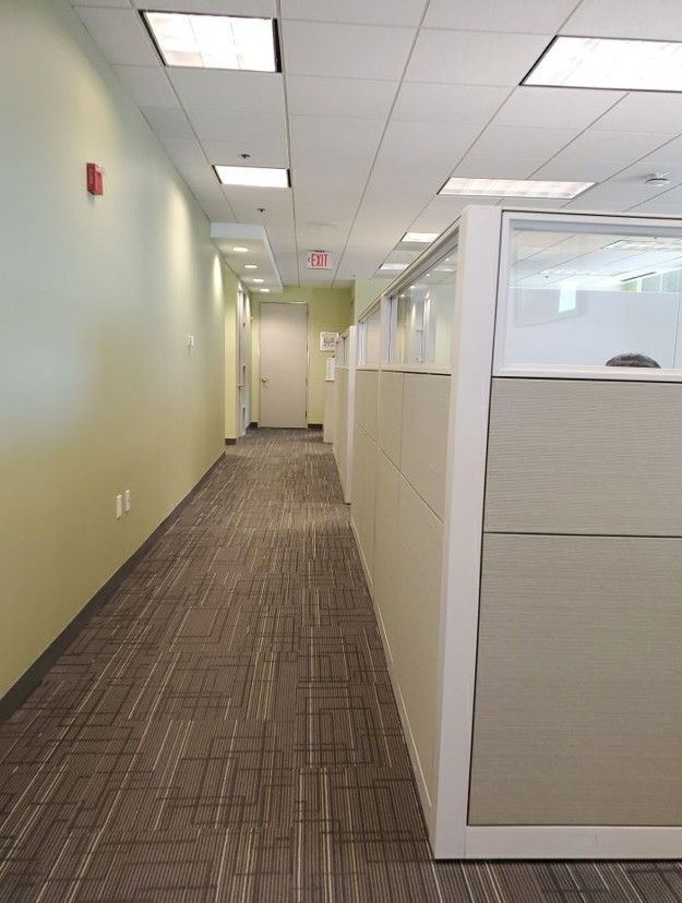 Empty office hallway with beige walls, carpet, ceiling lights, and cubicle partitions on the right