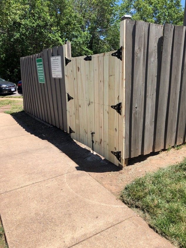 Wooden fence gate beside a sidewalk in a sunny outdoor area, with trees in the background