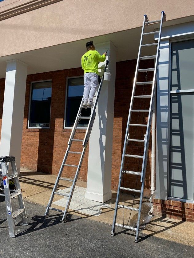 Worker on ladder painting a building’s white exterior trim under a covered walkway.