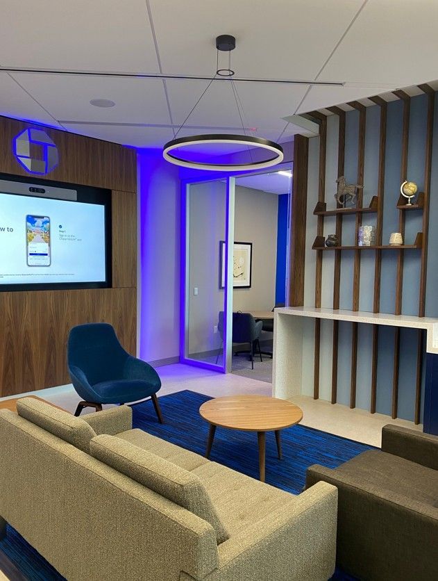 Modern office lounge with sofas, round table, blue accent lighting, wall screen, and decorative shelves