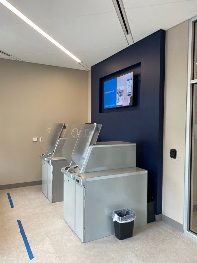 Two self-service kiosk machines in a hallway with a wall-mounted screen and a trash bin nearby.