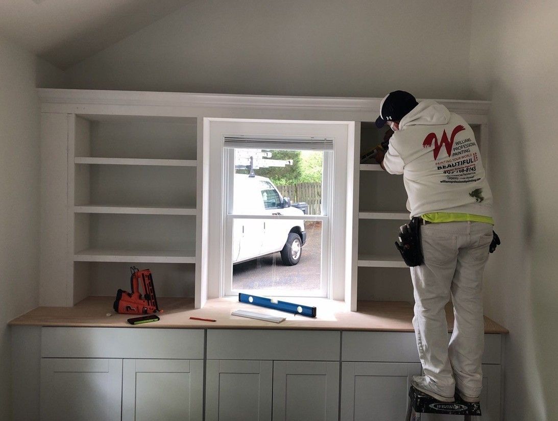Worker installing trim around a window above built-in cabinets in a bright room