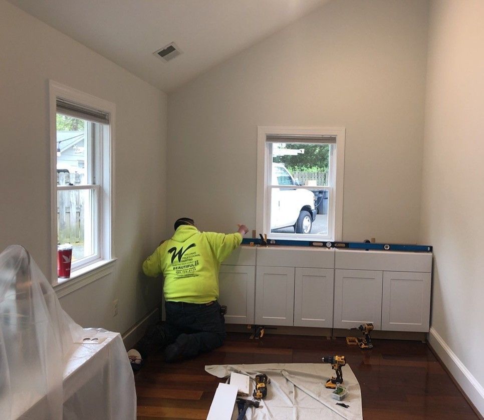 Worker installing cabinets in a bright, empty room with white walls and two windows.