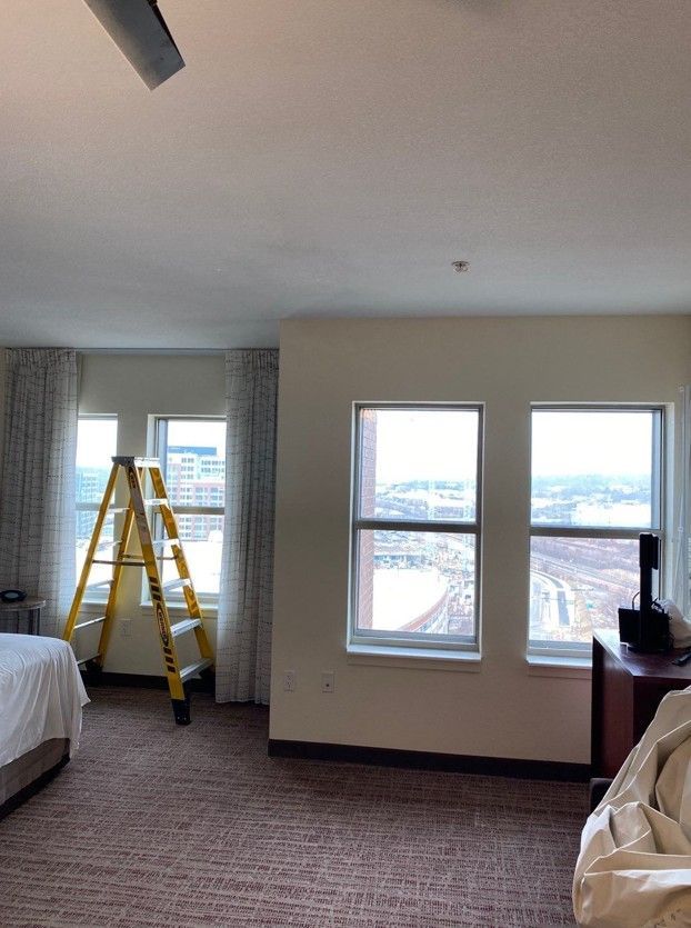 Hotel room interior with two windows, a yellow ladder by the curtain, and a bed partially visible.