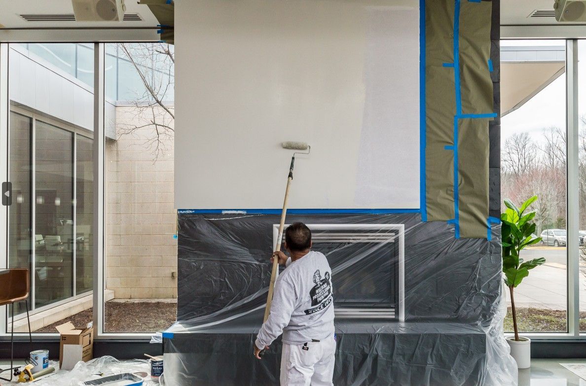Person painting a tall wall indoors, with windows and a protected fireplace area nearby.