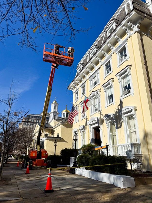 Orange boom lift beside a pale yellow building with flags and a church under a blue sky