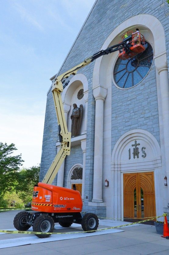 Orange cherry picker lift raises worker beside church facade with arched windows and wooden doors