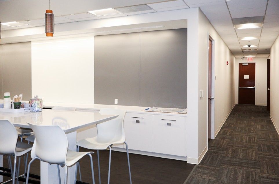 Office break room with white tables, chairs, cabinets, and a carpeted hallway leading to a closed door.