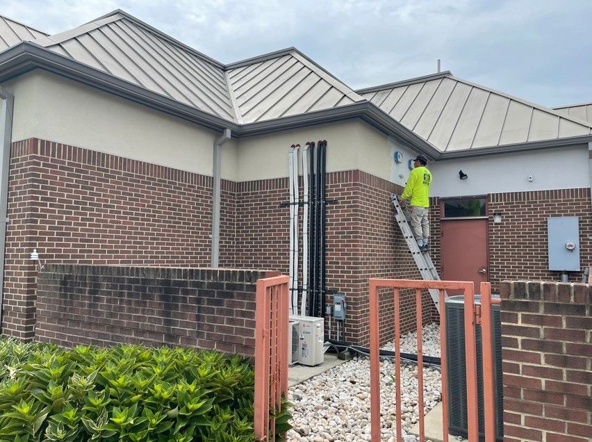 Worker on ladder inspecting a brick house roof and exterior wall near a gated entrance